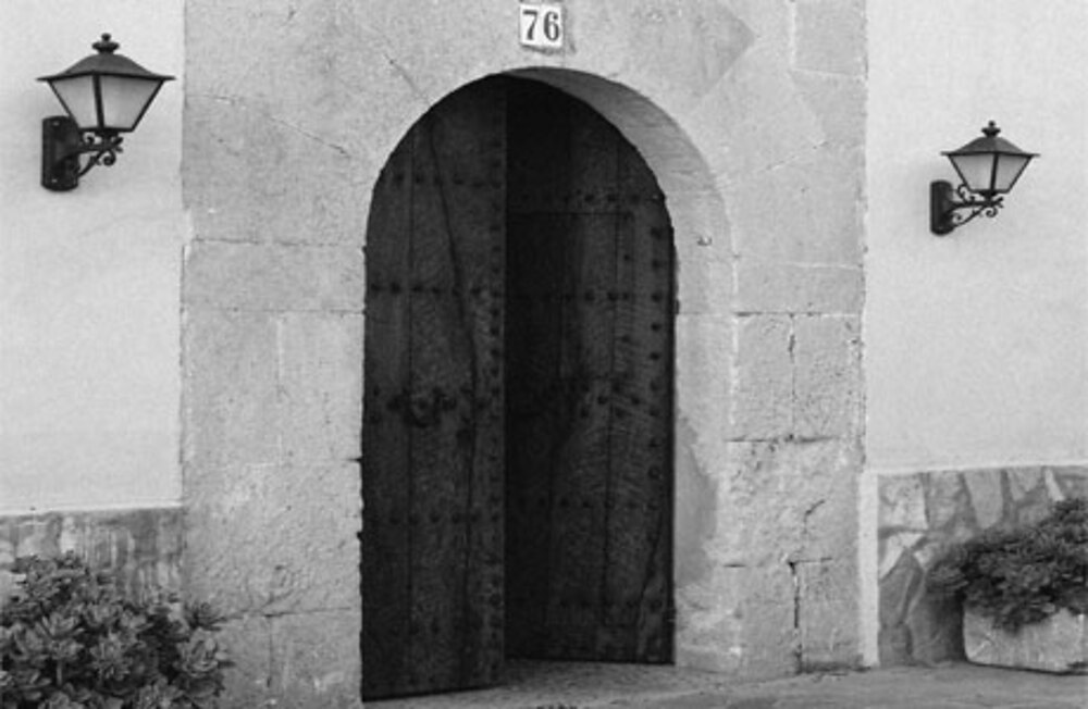 Black-and-white detail of the studded arched wooden door of Son Terrassa, numbered 76, between two wrought-iron wall lanterns