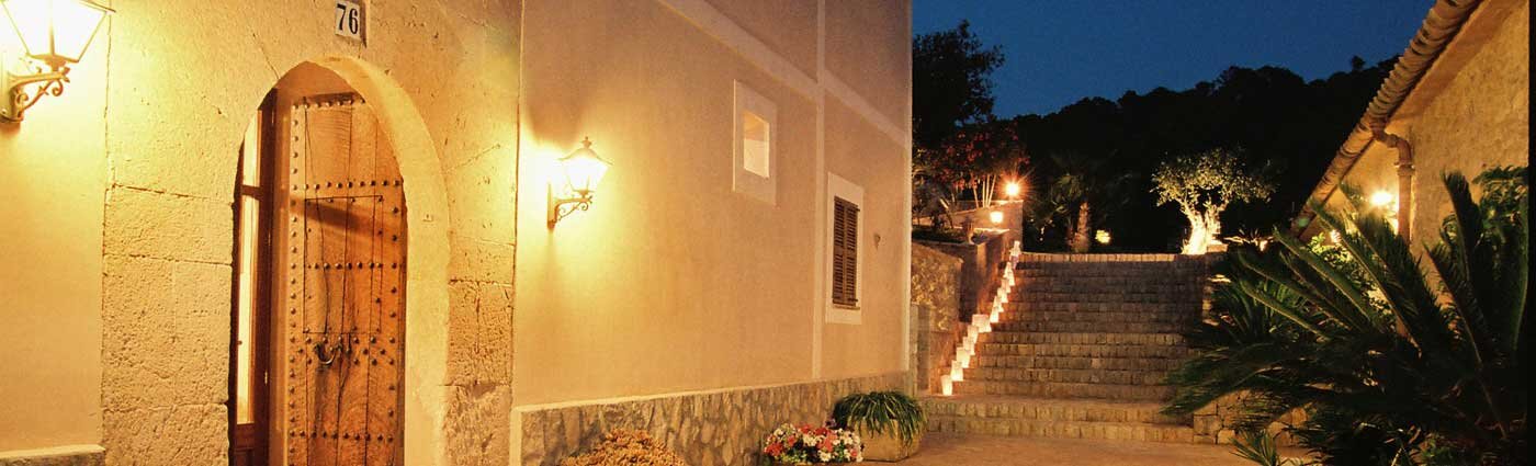 The honey-coloured marès facade of Son Terrassa at blue hour: the iron-studded arched doorway numbered 76 lit by wrought-iron lanterns, beside warm-lit stone garden steps rising past olive trees