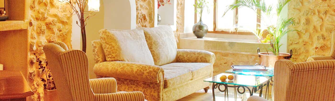 The sitting room at Son Terrassa: exposed marès stone walls, floral sofas, a fireplace and a sunlit window seat with lemons on a glass table