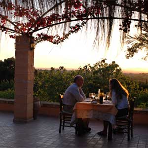 The terrace at Son Terrassa at sunset: a candlelit table for two under a pergola heavy with bougainvillea, the cultivated plain of Mallorca falling away to a far, hazy line of coast