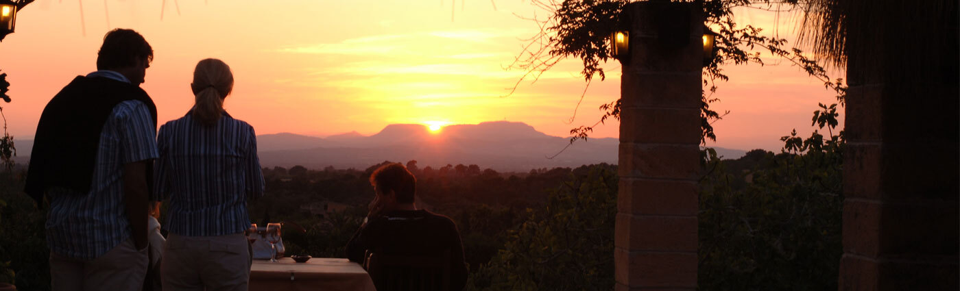 Two guests on the Son Terrassa terrace watching the sun set behind the mountains of Mallorca, between lantern-lit marès stone pillars